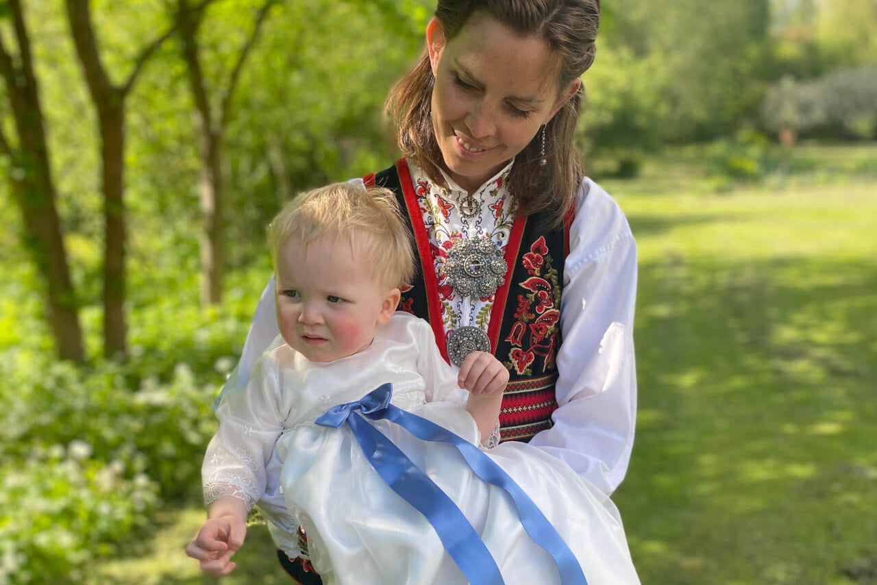 Woman in traditional outfit holding a child wearing a christening dress with a blue ribbon bow in a park