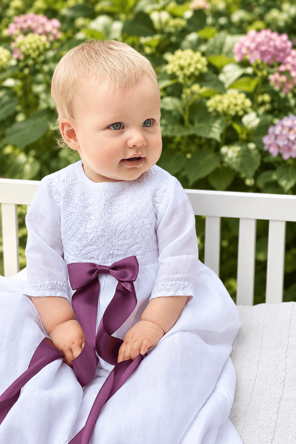 A baby girl wearing a white christening dress with a purple satin bow sits on a white bench in front of a garden background with green hydrangeas.