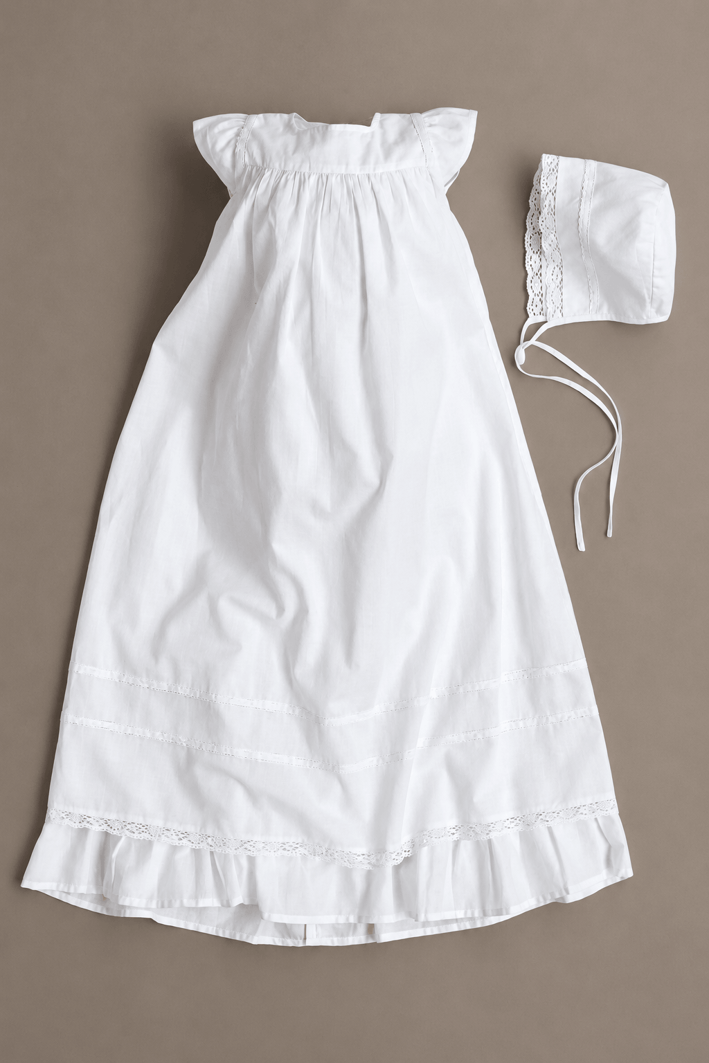 A white baby christening dress with ruffled shoulder straps and a matching bonnet with lace trim, laid flat on a neutral taupe background.