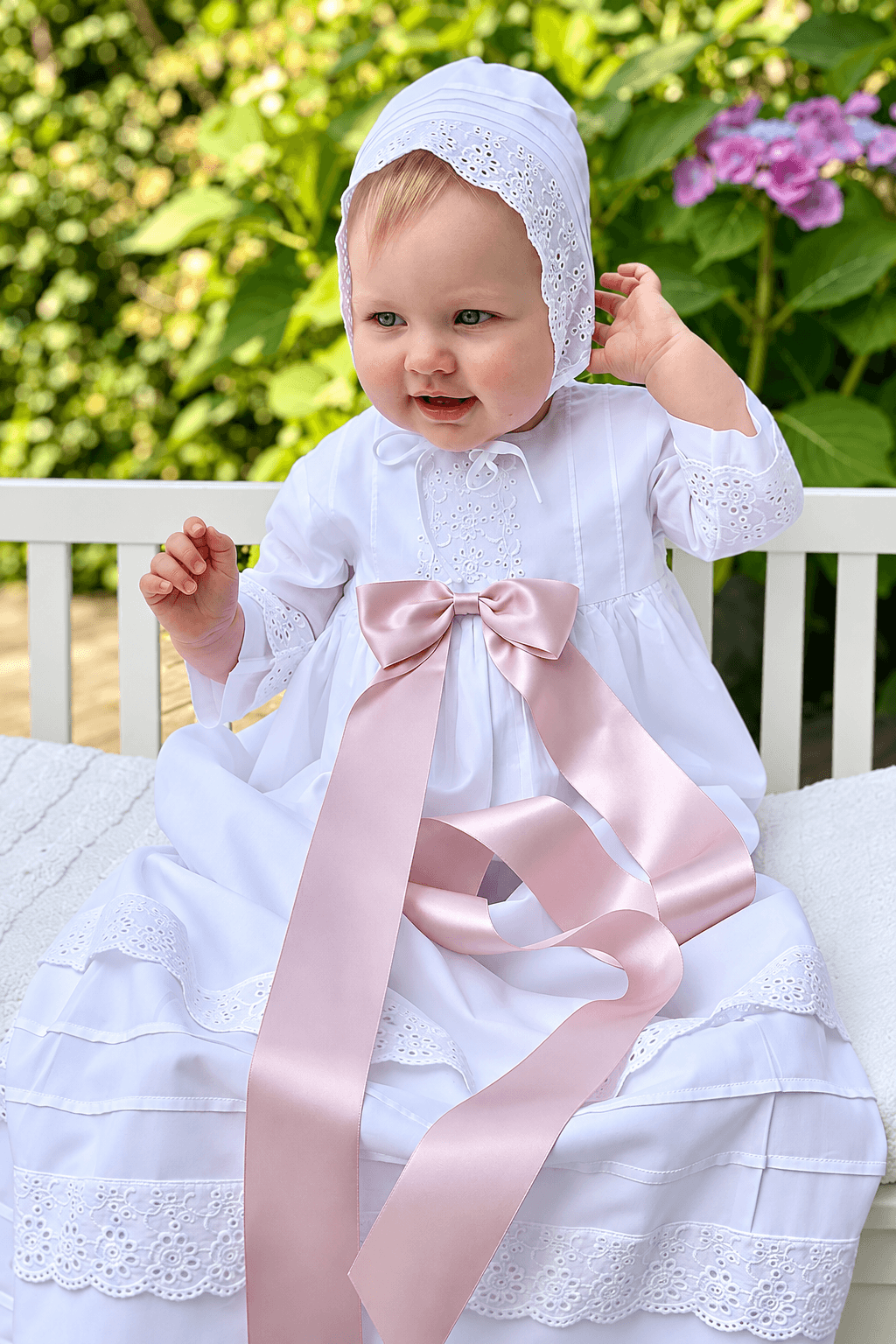 A baby wearing a white christening dress with a large pale pink satin bow and matching white bonnet, sitting outdoors on a white bench with green foliage in the background.