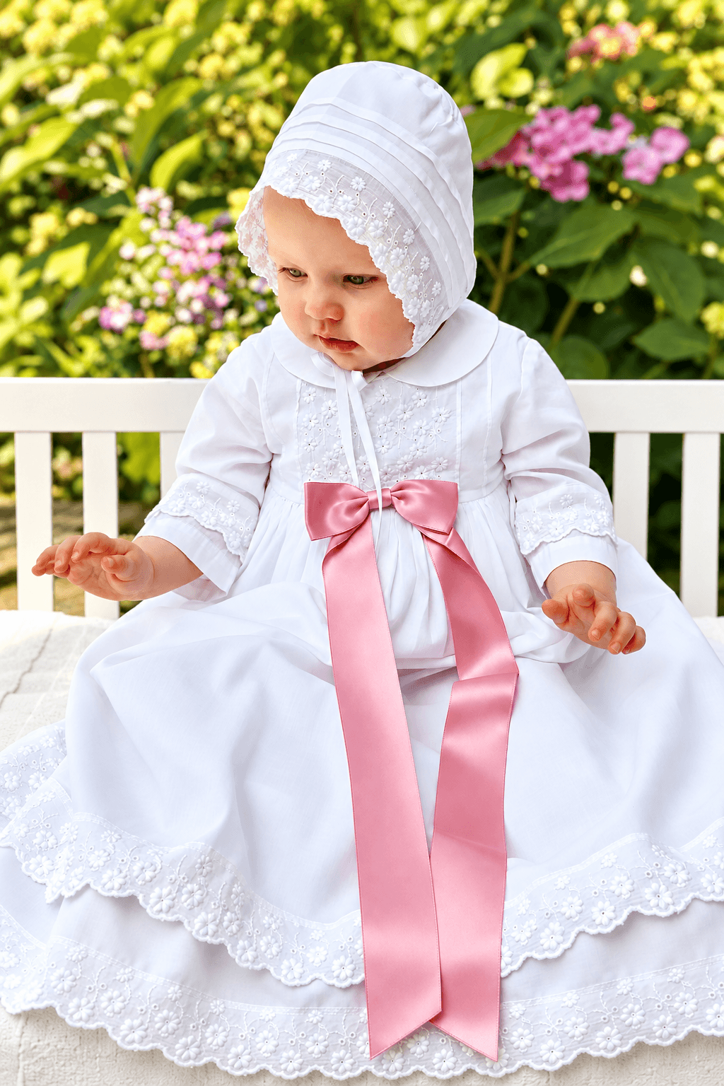 A baby wearing a white christening dress with a matching bonnet and a large pink satin bow, sitting on a white bench with a garden background.
