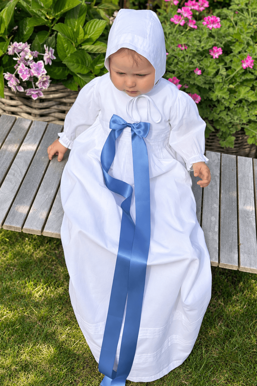 A baby wearing a white christening dress with a large blue ribbon bow, sitting on a wooden bench in a garden setting.